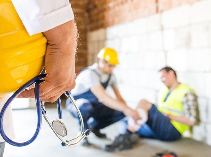 Profesional de salud ocupacional con casco y estetoscopio; al fondo, atención a una persona trabajadora lesionada